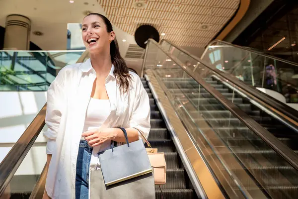 Esmer kadın yerel bir alışveriş merkezinde yürüyen merdivenle aşağı iniyor. Alışveriş torbaları taşıyor ve aldıkları kıyafetlerle mutlu.. 