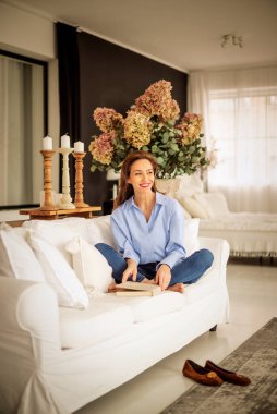 Güzel bir kadın evde rahatlatıcı bir günün tadını çıkarıyor. Orta yaşlı bir kadın rahat koltuğunda oturup kitap okurken gülümsüyor.