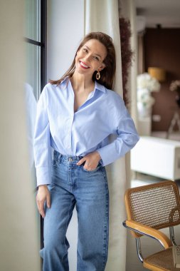 Evin penceresinde dinlenirken orta yaşlı, neşeli bir bayan gülümsüyor. Güzel kadın mavi gömlek ve kot pantolon giyiyor..  