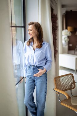 Evin penceresinde dinlenirken orta yaşlı, neşeli bir bayan gülümsüyor. Güzel kadın mavi gömlek ve kot pantolon giyiyor..  