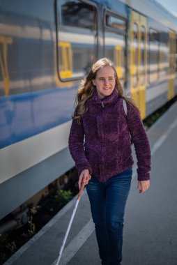 Şehrin dışındaki tren istasyonunda yürüyen beyaz bastonlu kör kadın portresi. Treni bekleyen görme engelli bir kadın. Günlük kıyafetler..  