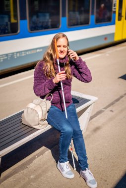 Beyaz bastonlu kör kadın portresi. Şehrin dışındaki tren istasyonunda bankta oturuyor. Treni bekleyen ve telefon kullanan görme engelli bir kadın. Günlük kıyafetler. Tam uzunluk.  