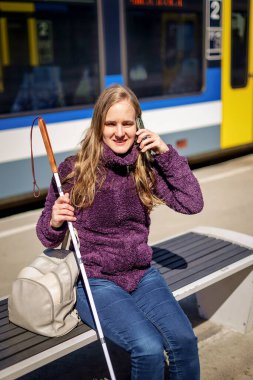 Beyaz bastonlu kör kadın portresi. Şehrin dışındaki tren istasyonunda bankta oturuyor. Treni bekleyen ve telefon kullanan görme engelli bir kadın. Günlük kıyafetler. Tam uzunluk.  