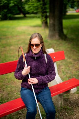 Beyaz baston tutan ve parktaki bankta oturan görme engelli bir kadın. Gülümseyen kadın rahat ceket ve kot pantolon giyiyor..  