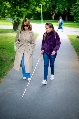 Şehir parkında kadın arkadaşıyla yürüyen görme engelli bir kadın. Görme engelli arkadaşına yardım eden olgun, beyaz bir kadın. Tam uzunluk.  