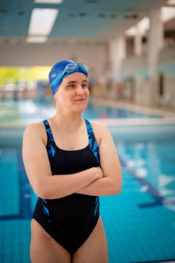 Kapalı bir yüzme havuzunda havuz kenarında duran görme engelli bir kadın. Mayo ve yüzme şapkası takan kendinden emin bir kadın..  