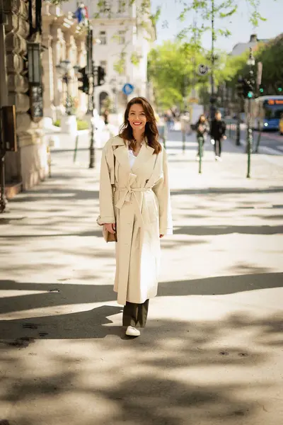 Güneşli bir günde, uzun saçlı, kendinden emin bir kadın şehir caddesinde yürüyor. Trençkot giyen ve neşeli gülümseyen çekici bir kadın. Tam boy çekim.  