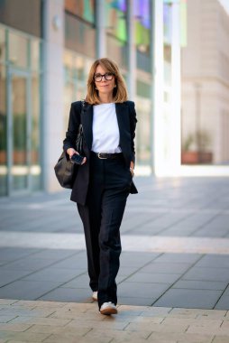 Ofis bölgesinde yürüyen orta yaşlı bir iş kadını. Sarı saçlı, siyah takım elbiseli ve gülümseyen bir kadın. Tam boy çekim.  