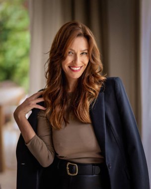 Rahat dairesinin penceresinde duran gülümseyen kadının portresi. Siyah ceket ve etek giyen orta yaşlı bir kadın.. 