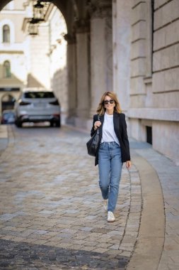 Olgun bir kadın bir sonbahar günü şehir caddesinde yürüyor. Siyah ceket ve kot giyen sarışın kadın. Tam boy çekim.  