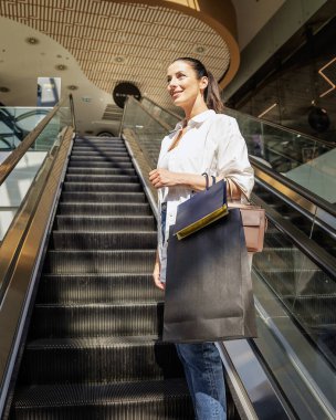 Esmer bir kadın yerel bir alışveriş merkezinde yürüyen merdivenle yukarı çıkıyor. Alışveriş torbaları taşıyor ve aldıkları kıyafetlerle mutlu.. 