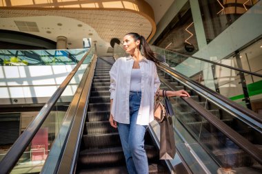 Esmer kadın yerel bir alışveriş merkezinde yürüyen merdivenle aşağı iniyor. Alışveriş torbaları taşıyor ve aldıkları kıyafetlerle mutlu.. 