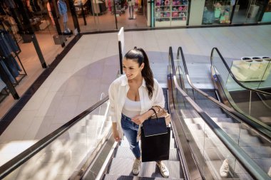 Esmer bir kadın yerel bir alışveriş merkezinde yürüyen merdivenle yukarı çıkıyor. Alışveriş torbaları taşıyor ve aldıkları kıyafetlerle mutlu.. 