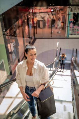 Esmer bir kadın yerel bir alışveriş merkezinde yürüyen merdivenle yukarı çıkıyor. Alışveriş torbaları taşıyor ve aldıkları kıyafetlerle mutlu.. 
