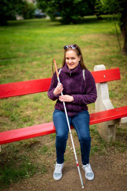 Beyaz baston tutan ve parktaki bankta oturan görme engelli bir kadın. Sıradan ceket ve kot pantolon giyen gülümseyen bir kadın. Tam uzunluk.  