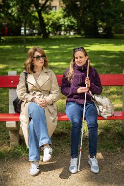 Şehir parkında kadın arkadaşıyla oturan görme engelli bir kadın. Görme engelli arkadaşına yardım eden olgun, beyaz bir kadın. Tam uzunluk.  