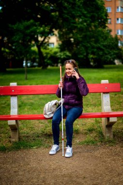 Beyaz baston tutan ve parktaki bankta oturan görme engelli bir kadın. Sıradan ceket ve kot pantolon giyen gülümseyen bir kadın. Tam uzunluk.  