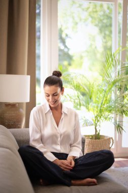 Orta yaşlı bir kadın pencerenin yanındaki kanepede oturuyor. Beyaz gömlek ve kot giyen esmer kadın. Tam uzunluk.