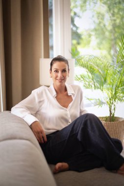 Orta yaşlı bir kadın pencerenin yanındaki kanepede oturuyor. Beyaz gömlek ve kot giyen esmer kadın. Tam uzunluk.