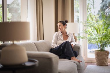 Orta yaşlı bir kadın pencerenin yanındaki kanepede oturuyor. Beyaz gömlek ve kot giyen esmer kadın. Tam uzunluk.