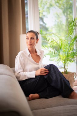 Orta yaşlı bir kadın pencerenin yanındaki kanepede oturuyor. Beyaz gömlek ve kot giyen esmer kadın. Tam uzunluk.