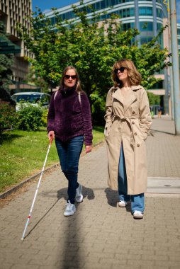 Görme engelli ve işitme engelli bir kadın, kız arkadaşıyla şehirde yürüyor. Görme engelli bir kadın elinde beyaz bir baston tutuyor ve konuşuyor. Tam uzunluk. 