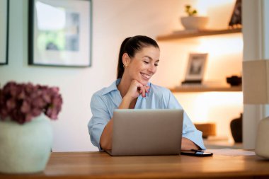 Orta yaşlı bir kadın bilgisayar kullanıyor ve evde çalışıyor. Masada oturan kendinden emin bir kadın ve neşeli bir gülümseme. Merkez Ofis. 