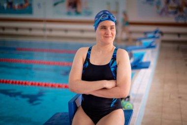 Kapalı bir yüzme havuzunda havuz kenarında oturan görme engelli bir kadın. Mayo ve yüzme şapkası takan kendinden emin bir kadın.. 