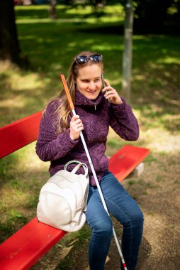 Beyaz baston tutan ve parktaki bankta oturan görme engelli bir kadın. Telefonunu kullanarak gülümseyen bir kadın. Tam uzunluk. 
