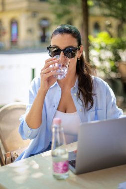 Masada oturmuş gülümseyen bir kadın, açık bir kafede dizüstü bilgisayarla çalışıyor. Sıradan kıyafetler giyen çekici bir kadın..