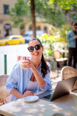 Masada oturmuş gülümseyen bir kadın, açık bir kafede dizüstü bilgisayarla çalışıyor. Sıradan kıyafetler giyen çekici bir kadın..