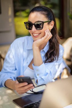 Masada oturmuş gülümseyen bir kadın, açık bir kafede dizüstü bilgisayarla çalışıyor. Sıradan kıyafetler giyen çekici bir kadın. Akıllı telefon kullanan kendine güvenen profesyonel bir kadın. . 