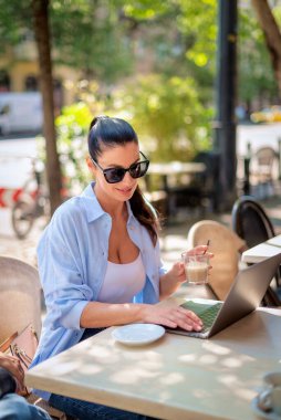 Masada oturmuş gülümseyen bir kadın, açık bir kafede dizüstü bilgisayarla çalışıyor. Sıradan kıyafetler giyen çekici bir kadın..
