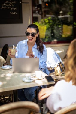 Masada oturmuş gülümseyen bir kadın, açık bir kafede dizüstü bilgisayarla çalışıyor. Sıradan kıyafetler giyen çekici bir kadın. Kendine güvenen profesyonel bir kadın. . 