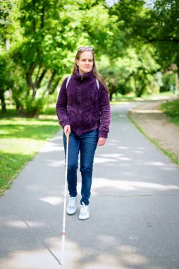 Beyaz bastonuyla parkta yürüyen görme engelli bir kadın. Sarı saçlı, sıradan kıyafetler giyen bir kadın. Tam boy çekim.