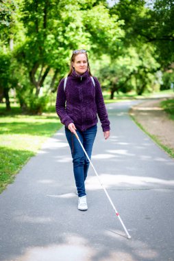 Beyaz bastonuyla parkta yürüyen görme engelli bir kadın. Sarı saçlı, sıradan kıyafetler giyen bir kadın. Tam boy çekim.