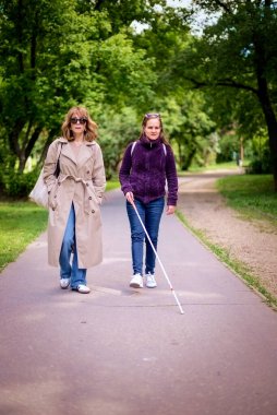 Şehir parkında kadın arkadaşıyla yürüyen görme engelli bir kadın. Görme engelli arkadaşına yardım eden olgun, beyaz bir kadın. Tam uzunluk. 
