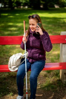 Beyaz baston tutan ve dışarıda akıllı telefon kullanan görme engelli bir kadın. Parktaki bankta oturan kendinden emin bir kadın.. 