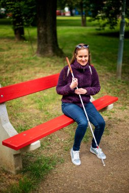 Beyaz baston tutan ve parktaki bankta oturan görme engelli bir kadın. Sıradan ceket ve kot pantolon giyen gülümseyen bir kadın. Tam boy çekim. 