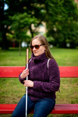 Beyaz baston tutan ve parktaki bankta oturan görme engelli bir kadın. Gülümseyen kadın rahat ceket ve kot pantolon giyiyor.. 