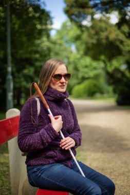 Beyaz baston tutan ve parktaki bankta oturan görme engelli bir kadın. Gülümseyen kadın rahat ceket ve kot pantolon giyiyor.. 