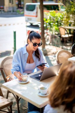 Masada oturmuş, dışarıda bir kafede dizüstü bilgisayarla çalışan neşeli bir kadın. Çekici bir kadın günlük kıyafetler giyiyor ve elinde akıllı bir telefon tutuyor.. 