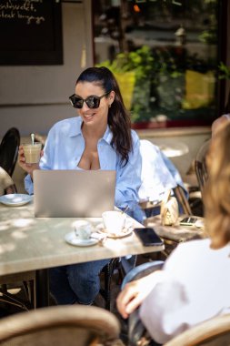 İki kadın kaldırımdaki kafede oturmuş konuşuyor. Bilgisayar laptopu kullanan esmer bir kadın. Gündelik kıyafetler ve güneş gözlüğü takan çekici bir kadın.. 