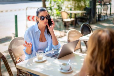 İki kadın kaldırımdaki kafede oturup konuşuyor. Bilgisayar bilgisayarı ve akıllı telefon kullanan esmer bir kadın. Gündelik kıyafetler ve güneş gözlüğü takan çekici bir kadın.. 
