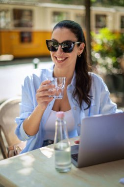 Masada oturmuş gülümseyen bir kadın, açık bir kafede dizüstü bilgisayarla çalışıyor. Sıradan kıyafetler giyen çekici bir kadın. Kendine güvenen profesyonel kadın neşeli gülümseme..