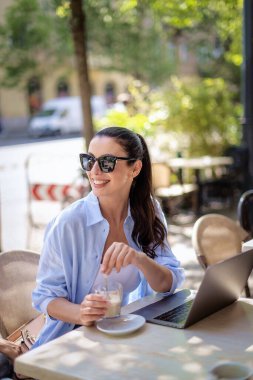 Masada oturmuş gülümseyen bir kadın, açık bir kafede dizüstü bilgisayarla çalışıyor. Sıradan kıyafetler giyen çekici bir kadın. Kendine güvenen profesyonel kadın neşeli gülümseme..