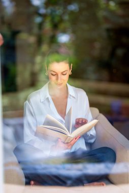 Güzel bir kadın evde rahatlatıcı bir günün tadını çıkarıyor. Orta yaşlı bir kadın rahat koltuğunda oturup kitap okurken gülümsüyor. Tam boy. Pencereden vurulmuş..