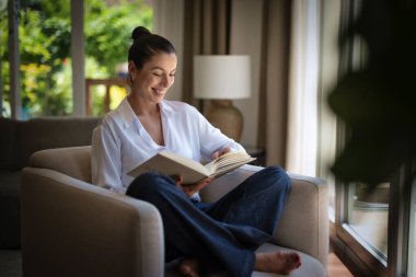Güzel bir kadın evde rahatlatıcı bir günün tadını çıkarıyor. Orta yaşlı bir kadın rahat koltuğunda oturup kitap okurken gülümsüyor. Tam uzunluk.