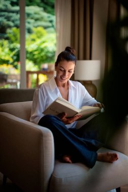 Güzel bir kadın evde rahatlatıcı bir günün tadını çıkarıyor. Orta yaşlı bir kadın rahat koltuğunda oturup kitap okurken gülümsüyor. Tam uzunluk.