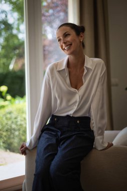 Modern evindeki koltukta rahatlayan esmer bir kadının portresi. Beyaz gömlek ve kot pantolon giyen beyaz kadın.. 